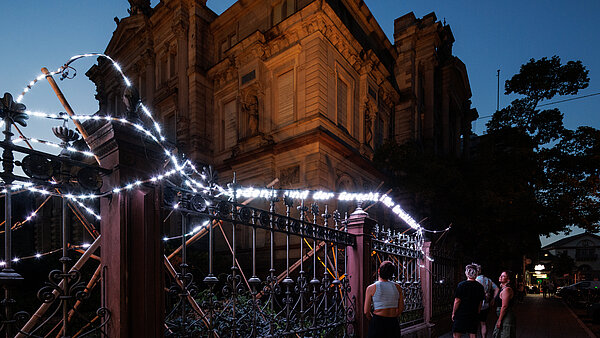 Ein Lichterband an einem Zaun vor einem historischen Gebäude, auf dem Worte gezeigt werden