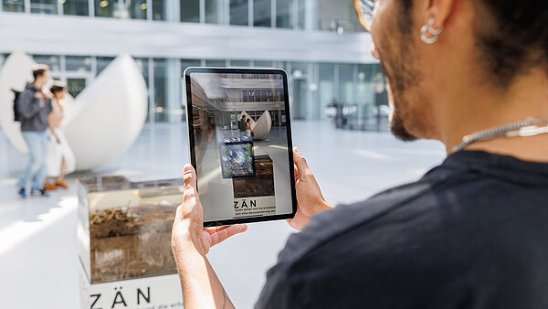 Eine Person in einem weißen Lichthof betrachtet durch die Kamera seines Tablets ein AR-Kunstwerk