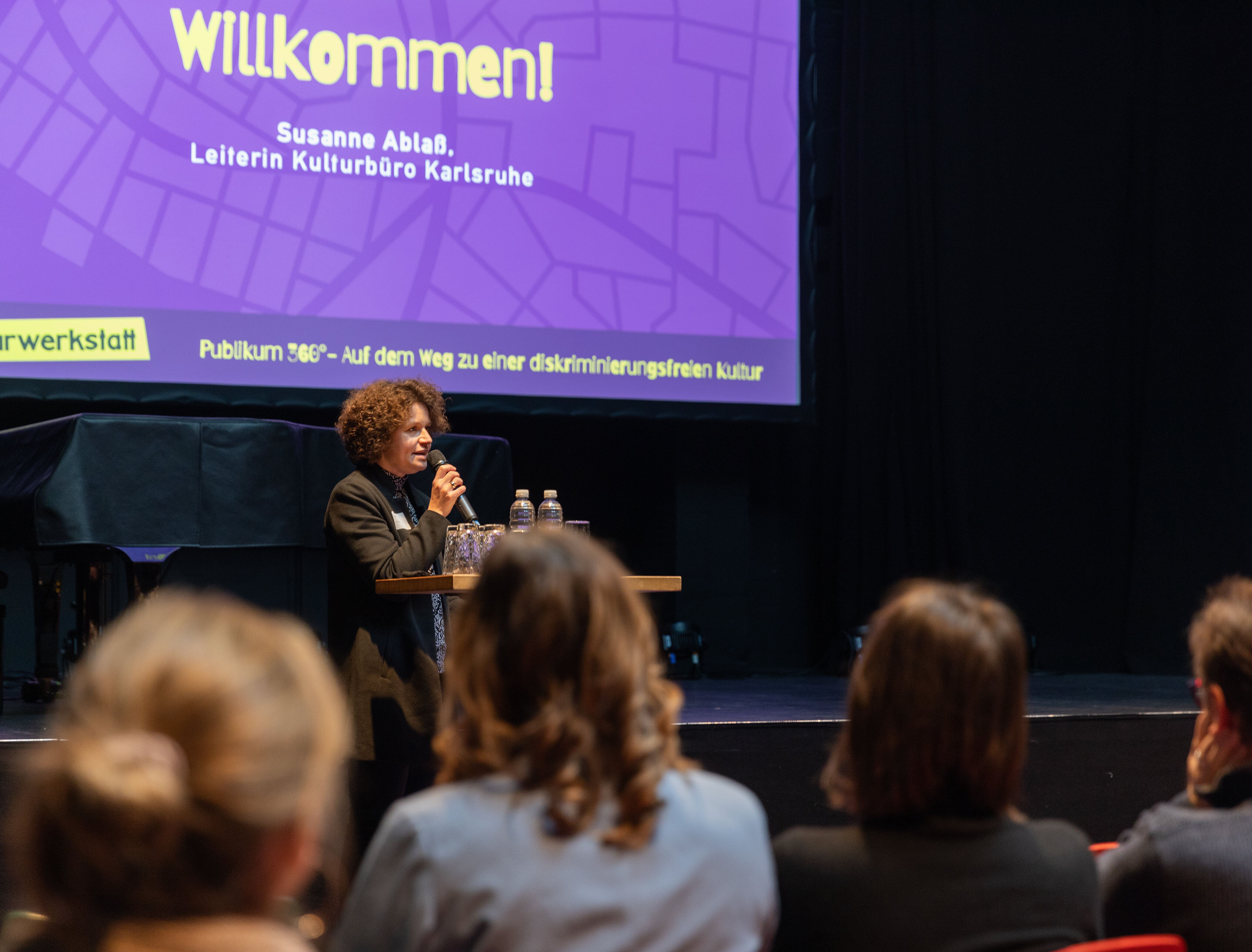 Leiterin des Kulturbüros steht vor Publikum auf der Bühne und begrüßt das Publikum