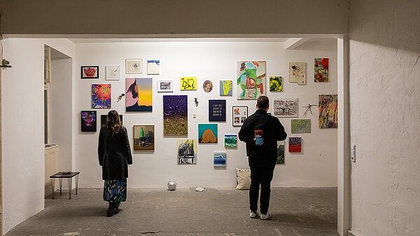 2 Personen vor einer Wand, die mit vielen Bildern behängt ist in einem leeren weißen Raum