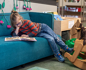 Lesendes Kind auf einem blauen Sofa neben einem Holzschaukelpferd.