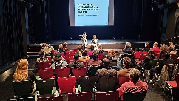 Bestuhlter Saal im Kulturzentrum Tollhaus mit Teilnehmenden, die sitzend der Einführung zum Runden Tisch Antirassismus / Antidiskriminierung am 15. November 2023 zuhören