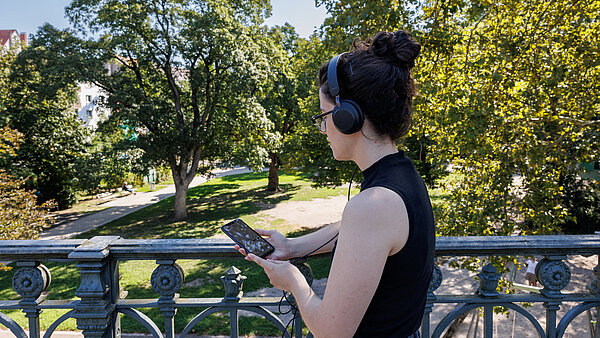 Eine Frau auf einer Brücke mit Kopfhörer und Smartphone in der Hand vor einer Park-Kulisse