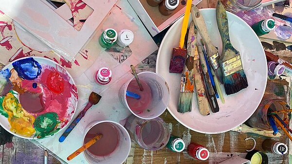 Blick von oben auf einen Tisch, der mit Matreialien zum Malen wie Pinsel und Farbbecher bedeckt ist.