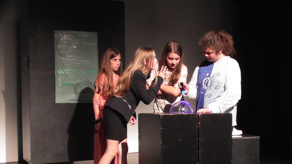 Eine Gruppe Kinder stehen im Halbkreis auf einer dunklen Bühne und schauen sich an