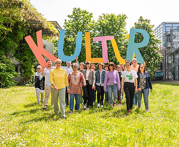 Eine Gruppe von Menschen an einem sonnigen Tag auf einer Wiese halten große Buchstaben, die das Wort Kultur bilden empor