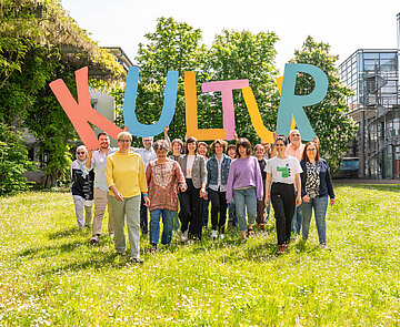 Eine Gruppe von Menschen an einem sonnigen Tag auf einer Wiese halten große Buchstaben, die das Wort Kultur bilden empor