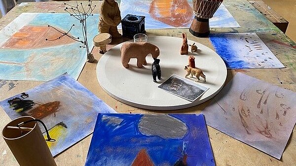 Blick von oben auf einen Holztisch, auf dem gemalte Bilder aus Pastell und Tierfiguren liegen
