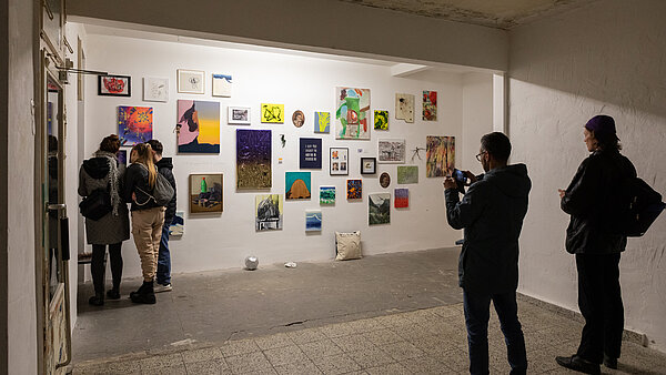 Eine Wand, die mit vielen Bildern behängt ist und fünf betrachtenden Personen davor