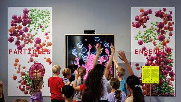 Kinder in Bewegung vor einem Bildschirm, auf dem deren Umrisse zu sehen sind und der daneben befindlichen Beschriftung Particles Ensemble