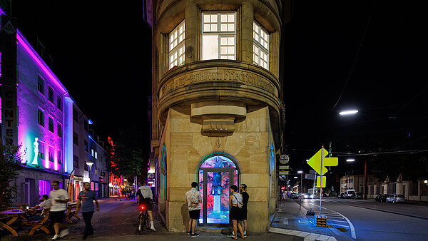 Personen nachts vor einem schmalen Gebäude, die ein Kunstwerk im Erdgeschoss betrachten